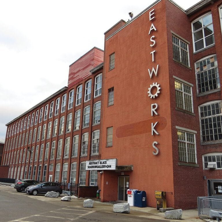 Rust-colored brick building with large windows, "EAST WORKS" vertical sign, and gallery signage.