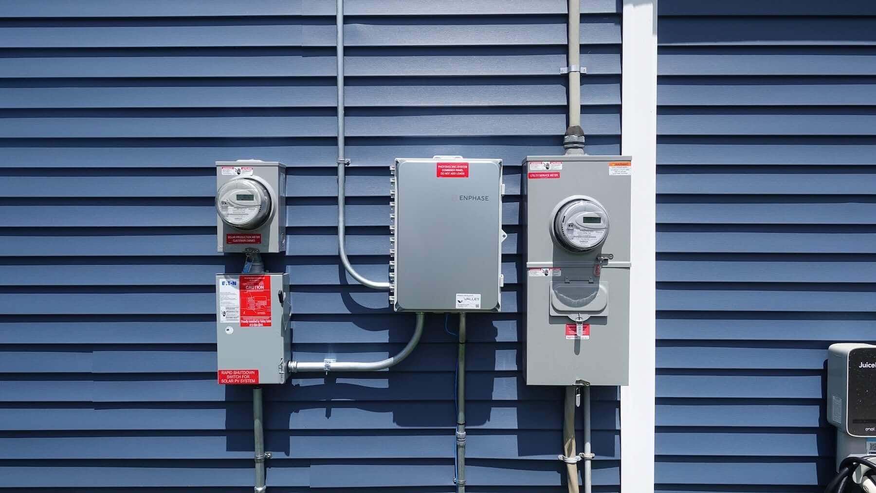 Electrical meters and solar power system components installed on a blue house siding.